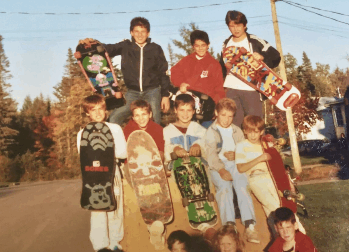 A group of boys pose outdoors on a street, holding colorful skateboards and smiling. Some are sitting, some standing, with trees and power lines visible in the background. The scene appears to be from the 1980s or 1990s.