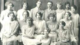 A vintage black-and-white group portrait of fifteen young adults from the early 20th century, including two men in suits and thirteen women in light-colored dresses, all seated or standing and facing the camera.