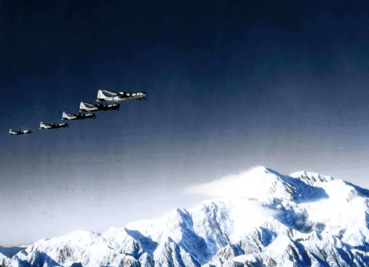 A formation of military aircraft flies in the sky above a snow-covered mountain range under a clear, deep blue sky.
