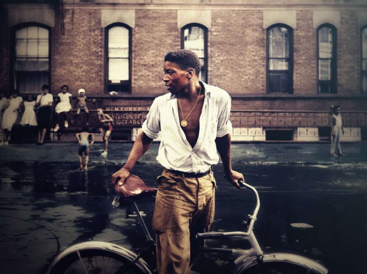 A young man in a white shirt and brown pants stands holding a bicycle on a city street, with a group of people and apartment buildings in the background.
