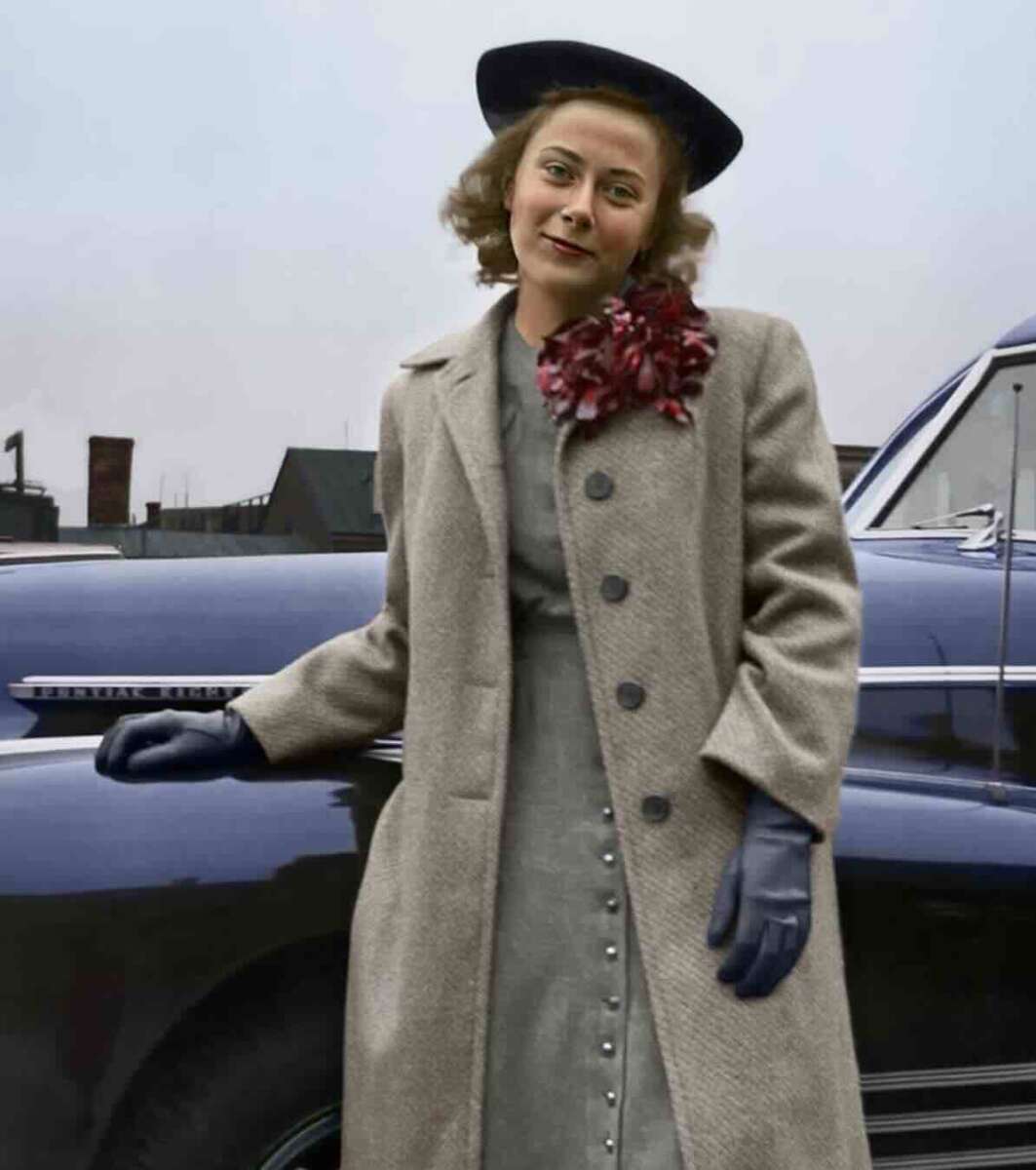 A woman in a beige coat, blue gloves, and a dark hat stands in front of a vintage navy blue car. She has a red flower pinned to her coat and is smiling slightly, with buildings visible in the background.