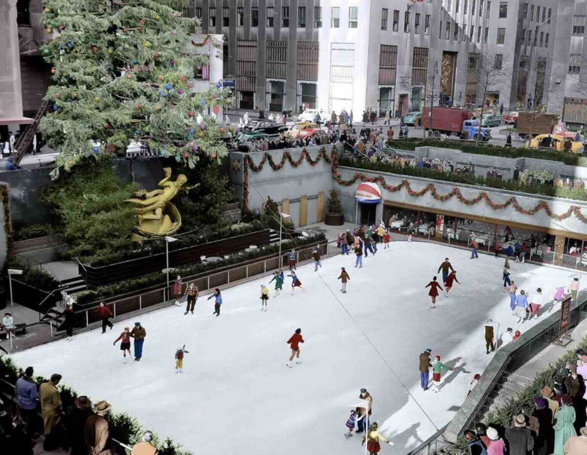 People ice skating at Rockefeller Center rink in New York City, surrounded by holiday decorations and a large Christmas tree, with golden statue and buildings in the background. Spectators watch from the sides.