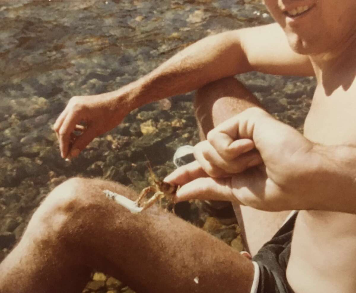 A shirtless person sitting by a rocky riverbank holds a small crayfish with their fingers, smiling. One arm rests on their knee, and water with visible rocks flows in the background.