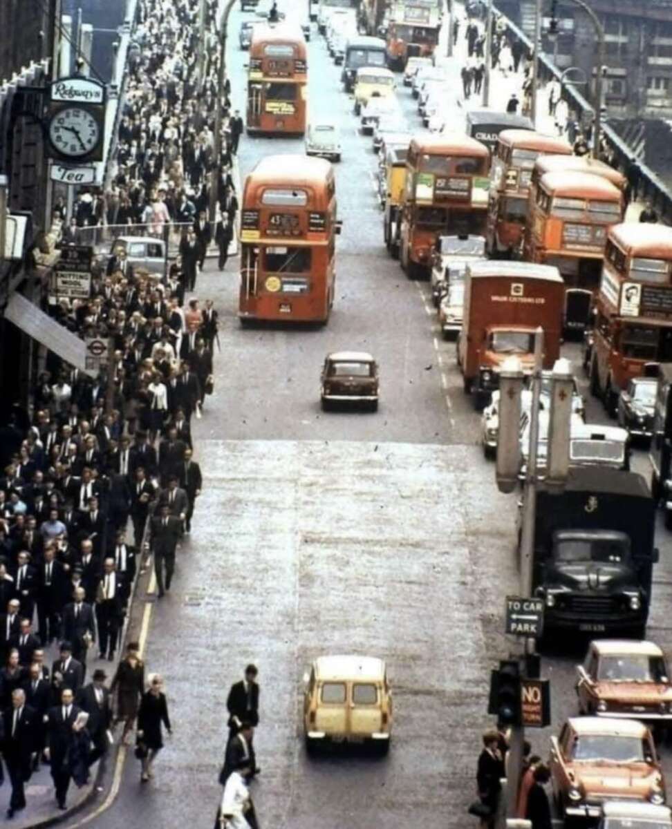 A busy city street in London, crowded with people on sidewalks and several red double-decker buses and cars on the road. The photo appears to be from the mid-20th century.