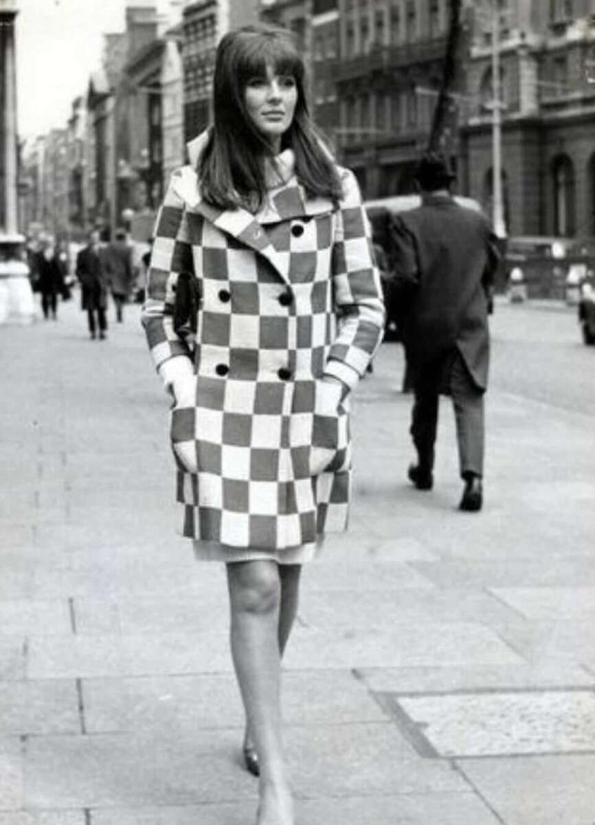 A woman walks confidently down a city street wearing a bold, checkered coat with her hands in the pockets. The photo is black and white, suggesting a vintage or 1960s fashion scene.