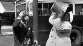 A black and white photo shows two men on a street; one wipes his forehead while the other drinks from a large metal container. A bus is visible in the background.