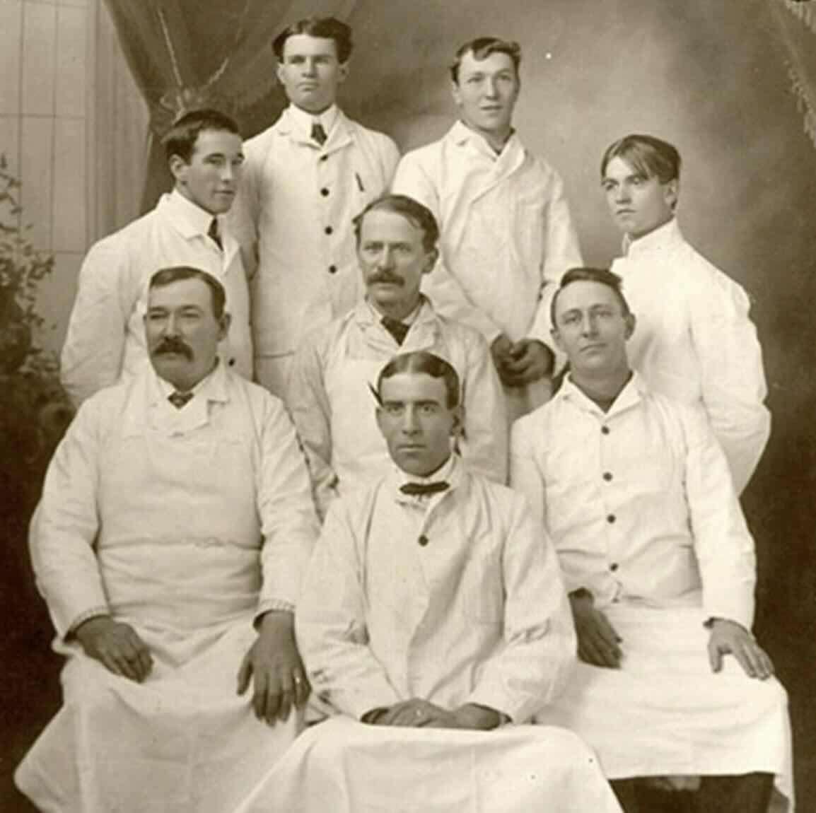 Seven men and one young man pose for a formal vintage photograph, all wearing white coats or uniforms, seated and standing in two rows against a studio backdrop.