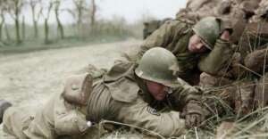 Two soldiers in green World War II uniforms and helmets lie on the ground beside a stone wall, one writing while the other rests his head on his arm. The scene is outdoors near a dirt road and trees.