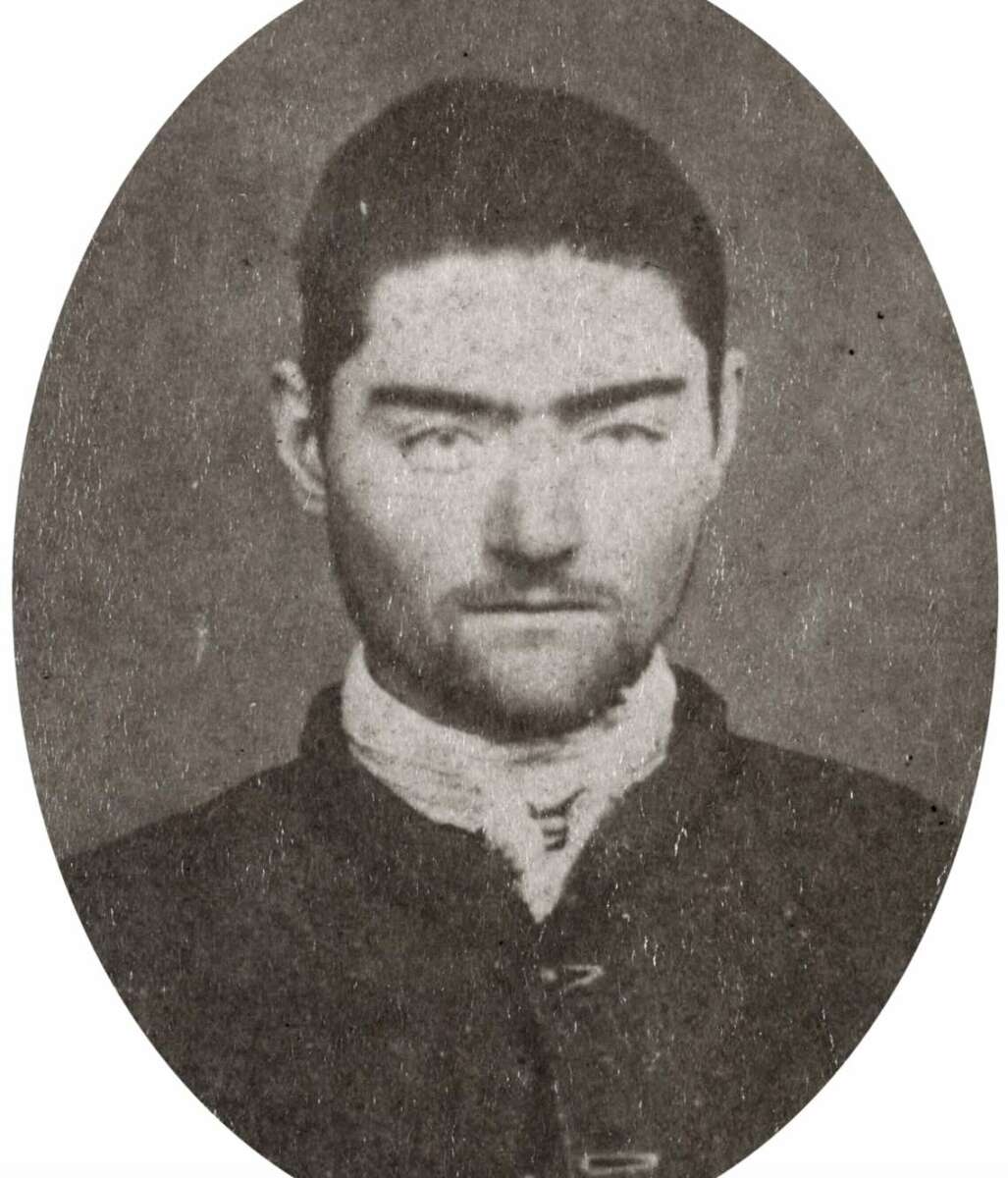 A black-and-white, oval-framed portrait of a young man with short dark hair, a trimmed beard, and strong eyebrows, wearing a high-collared shirt and dark buttoned jacket, looking seriously at the camera.