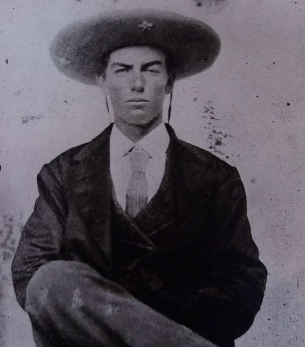 A black-and-white vintage photograph of a seated young man wearing a wide-brimmed hat, a suit jacket, waistcoat, and tie. He has a serious expression and sits with one leg crossed over the other.