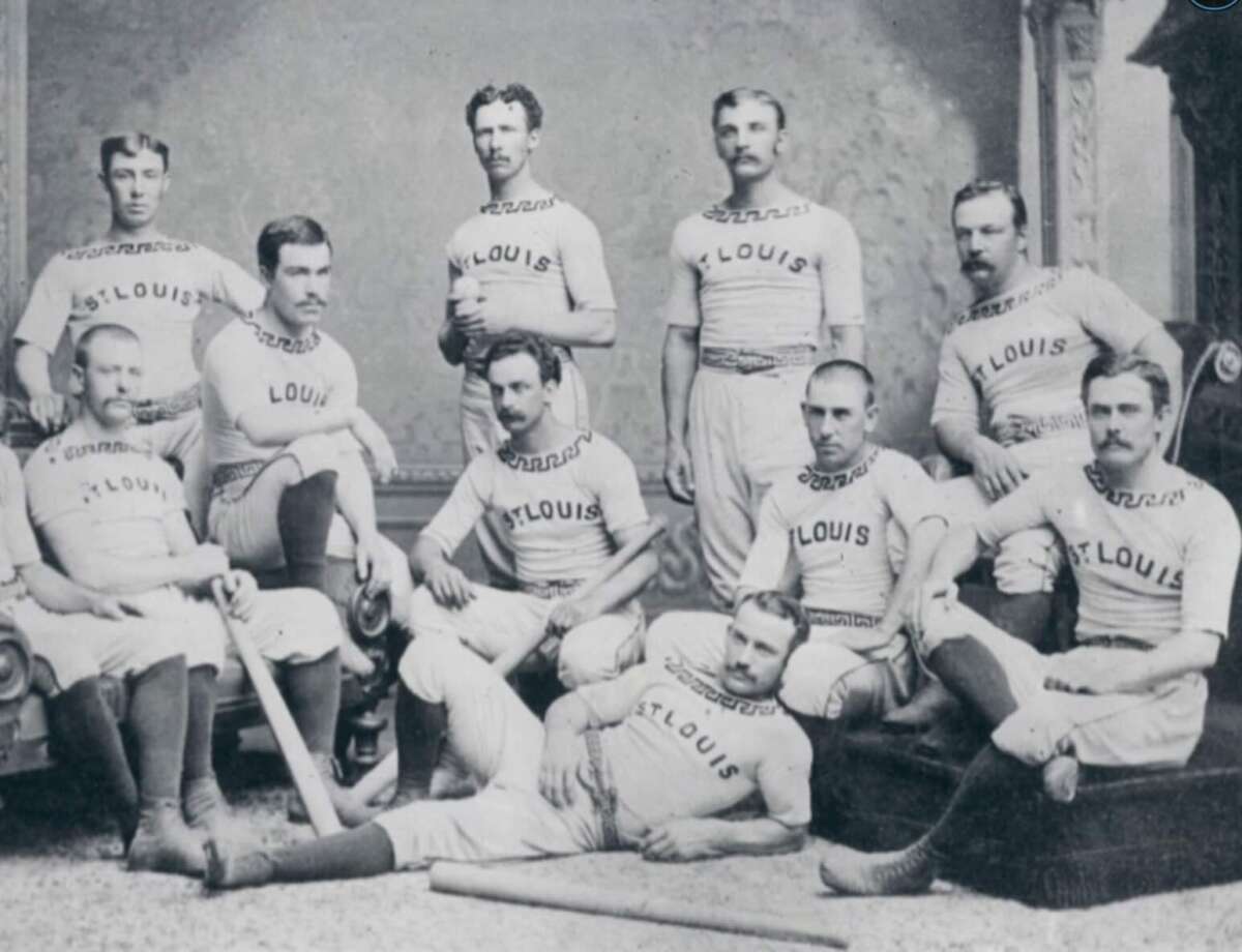 A vintage black-and-white photo of a baseball team, with ten men in matching "ST. LOUIS" uniforms posing together indoors; some standing, others seated or reclining with bats.