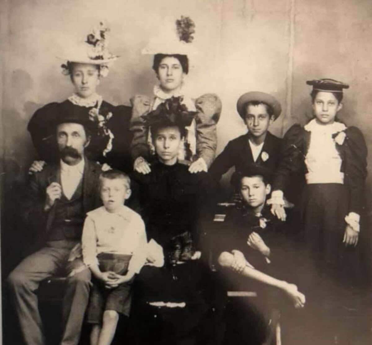 A sepia-toned vintage photograph of a family with eight members—four women in hats and dresses standing and seated, a bearded man in a suit, and three boys, one sitting on the man's lap—posing for a formal portrait.