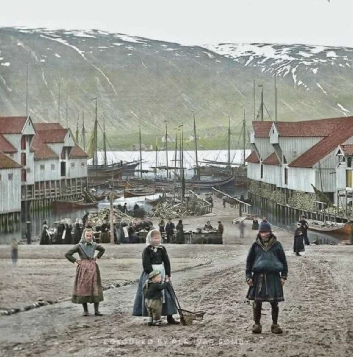 A historic, colorized photo of a Nordic fishing village. People in traditional clothing stand on a dirt road near wooden buildings and boats docked along a snowy, mountainous shoreline.