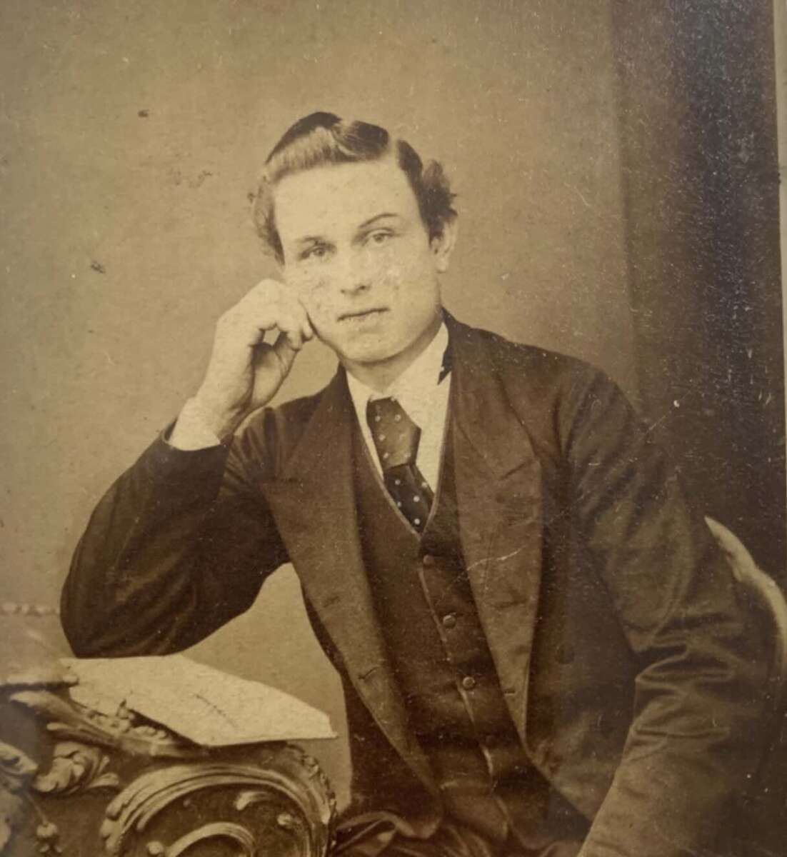 Young man in vintage formal attire sits with his elbow on an ornate table, resting his face on his hand. He looks directly at the camera, with old papers visible on the table beside him. The image is sepia-toned.