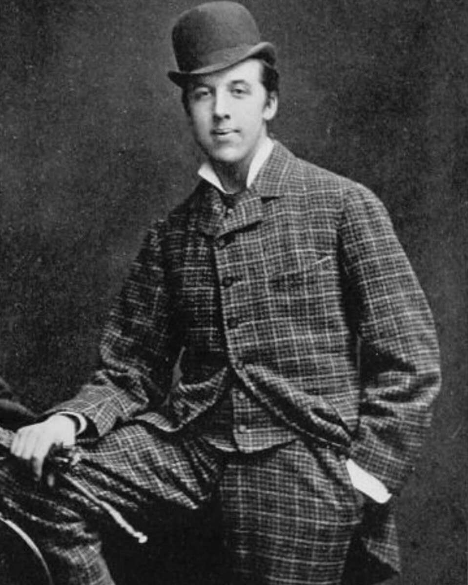 A young man in a plaid suit and bowler hat poses for a vintage black-and-white portrait, standing with one hand in his pocket and the other resting on a cane, against a dark studio backdrop.