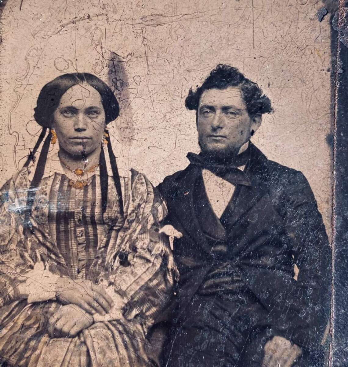 A vintage photograph of a seated woman and man in formal 19th-century clothing. The woman wears a striped dress and jewelry, while the man wears a dark suit and bow tie. Both have serious expressions, and the image is faded and scratched.