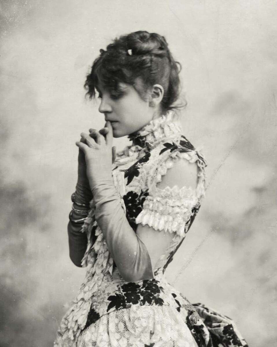 A vintage black-and-white photo of a young woman in profile, wearing an ornate dress with lace and floral patterns. She appears thoughtful, with her hands clasped near her face and her hair styled up.