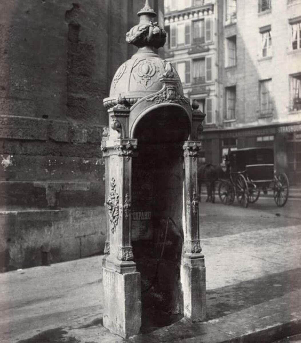 Ornate vintage drinking fountain with a domed roof and sculpted details stands on a city street; old buildings and a horse-drawn carriage are visible in the background.