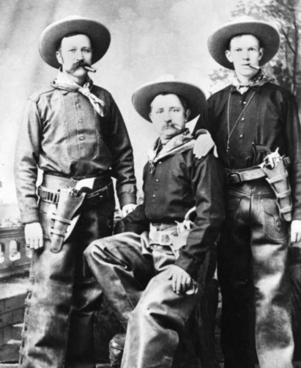 Three men dressed as cowboys, wearing wide-brimmed hats, neckerchiefs, and gun holsters, pose for a black-and-white photograph; two stand while one sits in the middle.