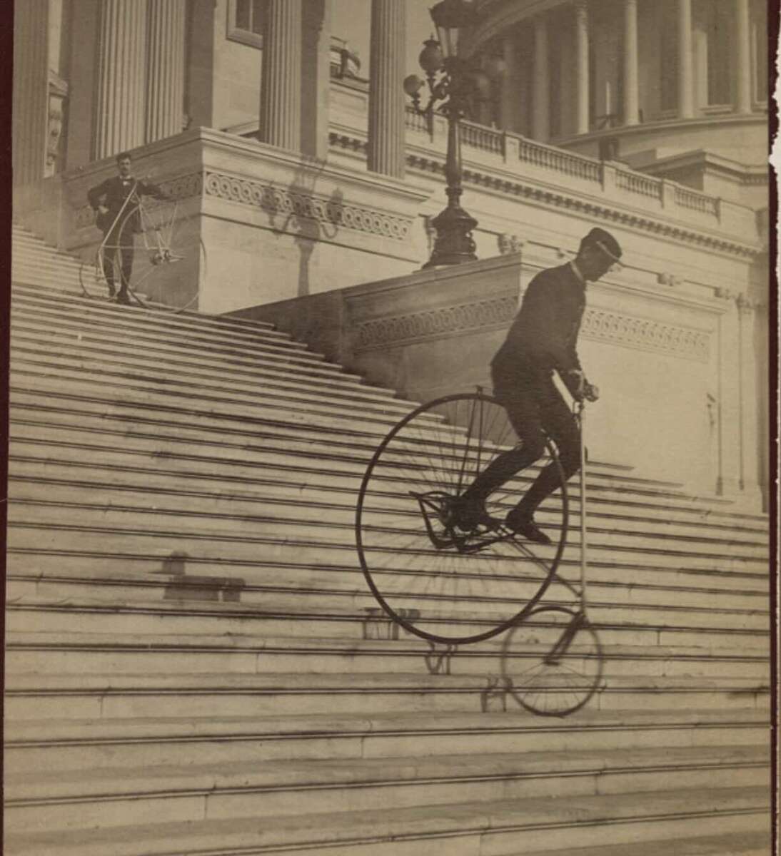 A man rides a penny-farthing bicycle down the steps of a large, classical building, while another man with a similar bicycle stands at the top of the stairs, watching.