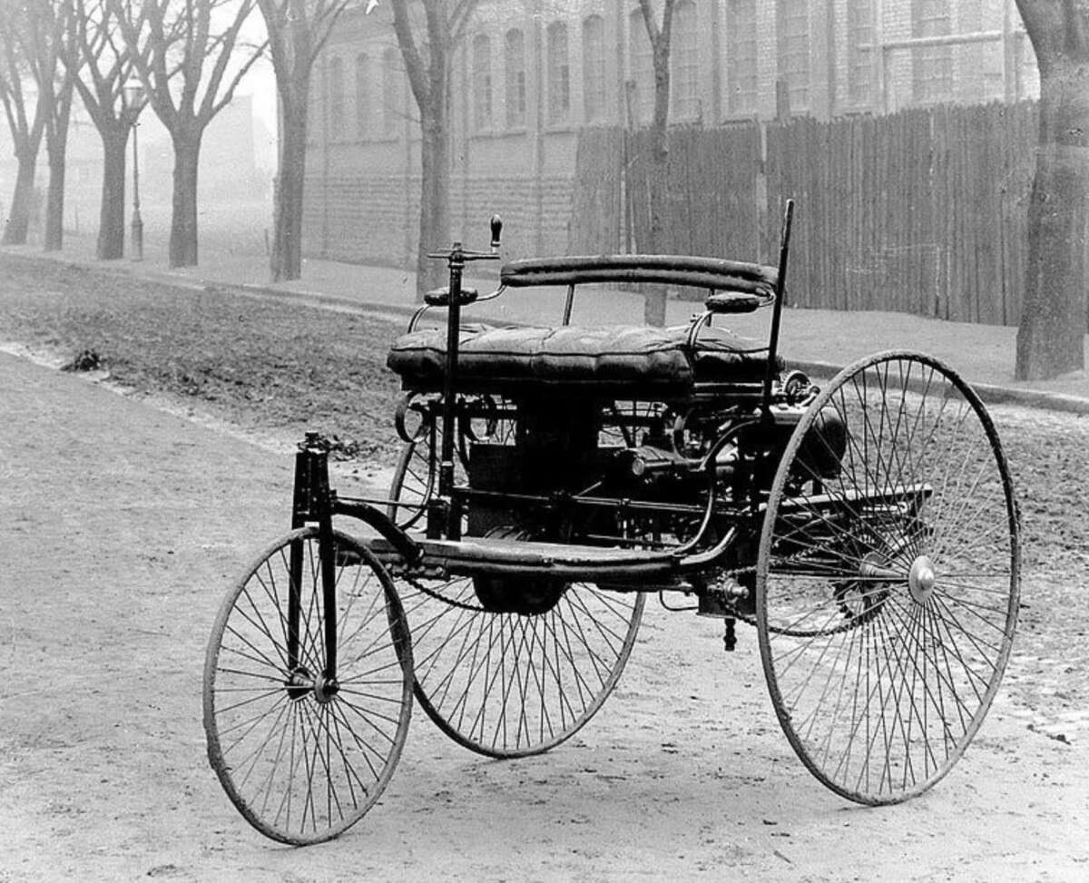 A black-and-white photo of an old-fashioned, three-wheeled motor vehicle with thin spoked wheels, parked on a dirt road lined with leafless trees and a tall wooden fence.