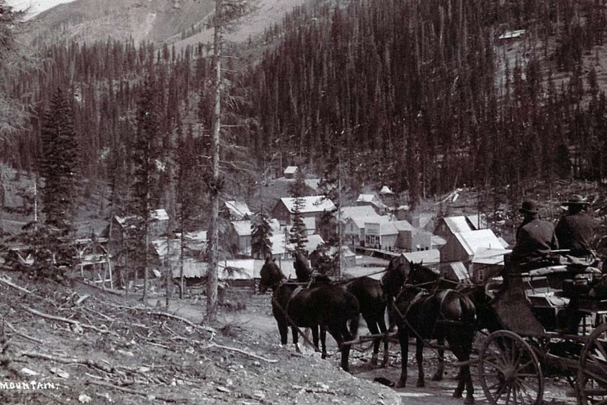A black-and-white photo shows a horse-drawn carriage on a dirt road overlooking a small mountain town with scattered wooden buildings surrounded by dense pine trees and hills.