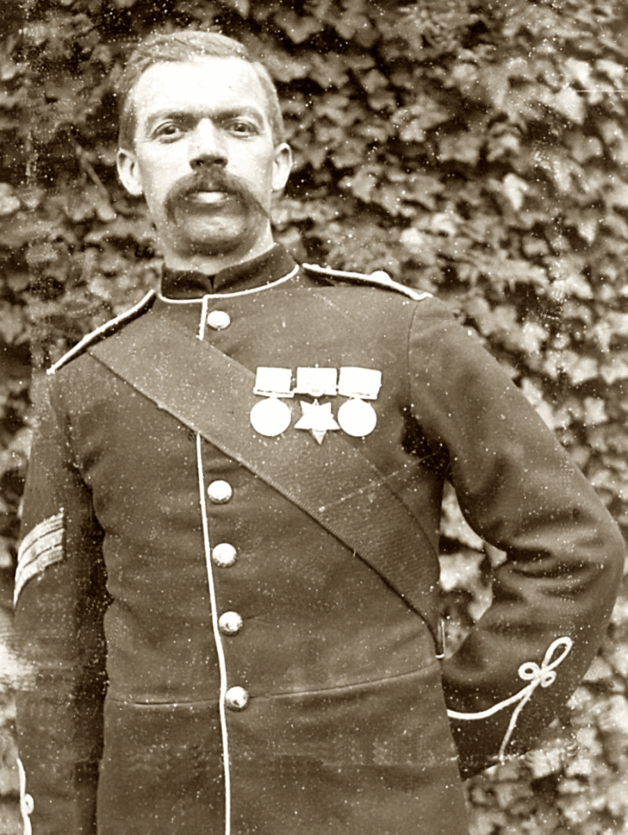 A man with a mustache wears a decorated military uniform with medals and a sash, standing in front of a leafy background.