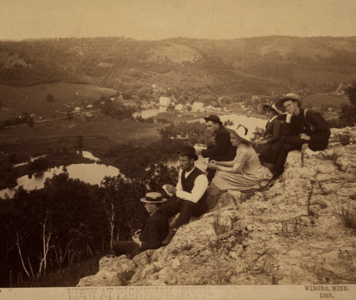 A group of men and women in 19th-century clothing sit on a rocky hilltop overlooking a scenic rural landscape with rivers, trees, and distant buildings in Winona, Minnesota, 1885.