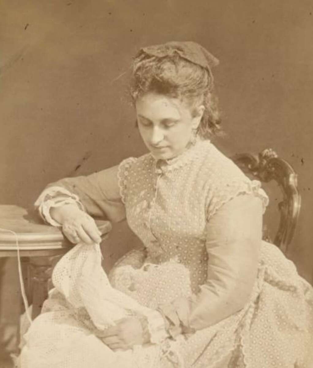 A sepia-toned photo of a woman in a 19th-century dress sitting on a chair, looking down as she works with needle and fabric, possibly sewing or embroidering, next to a small round table.
