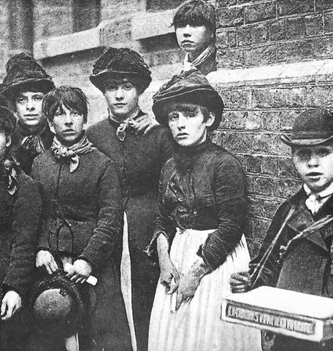 A group of young girls in old-fashioned clothing and hats stand against a brick wall; a boy in a cap and coat stands at the right holding a box with a strap. The photo appears to be from the late 19th or early 20th century.