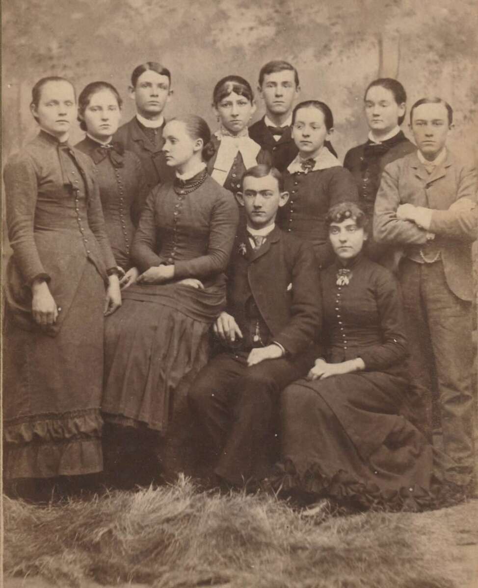 A sepia-toned vintage photo of eleven people, six women and five men, dressed in formal 19th-century clothing, posing together in a studio setting with a plain backdrop and straw on the floor.
