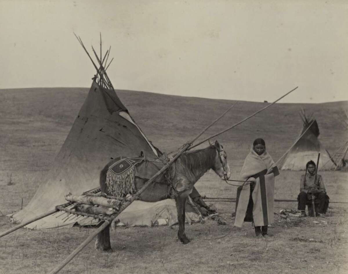 A person stands holding the reins of a horse carrying supplies and poles in front of a tipi, while another person sits nearby on the open plains. The scene appears historical and depicts Indigenous life.