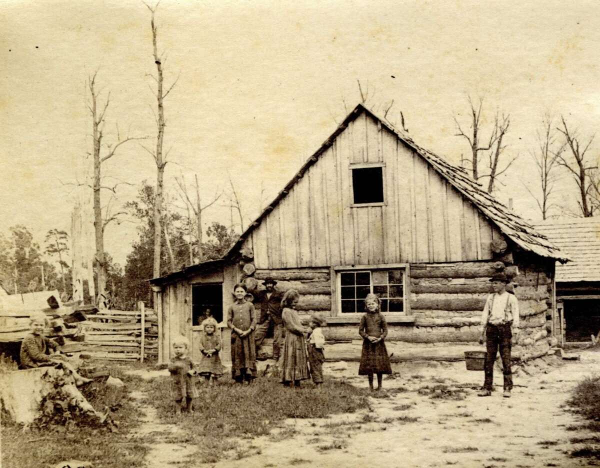A sepia-toned photo shows a family of eight, including children and adults, standing outside a rustic log cabin with a wooden fence and tall, leafless trees in the background.