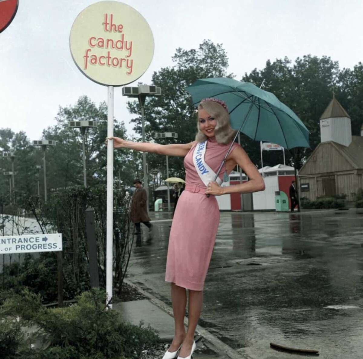 A woman in a pink dress with a sash reading "England" stands smiling under a teal umbrella by a sign that says "the candy factory," on a wet street with trees and buildings in the background.