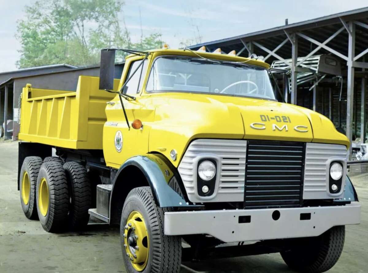 A bright yellow vintage CMC dump truck with six wheels is parked outdoors near a building with a metal roof. The truck has a distinctive, retro front grille and two black stripes on the fenders.