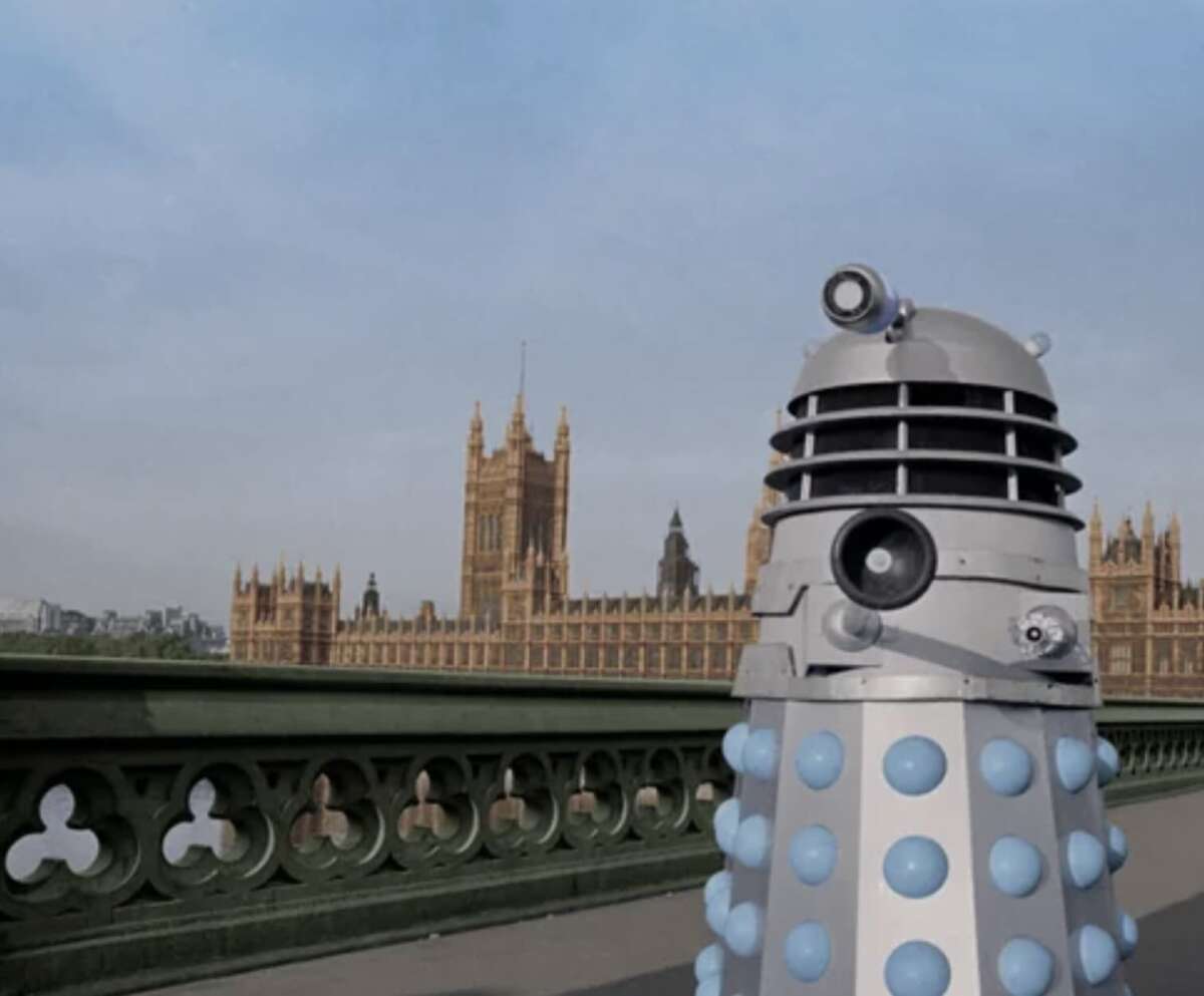 A Dalek robot from Doctor Who stands on a bridge with the Houses of Parliament and Big Ben in the background on a clear day in London.