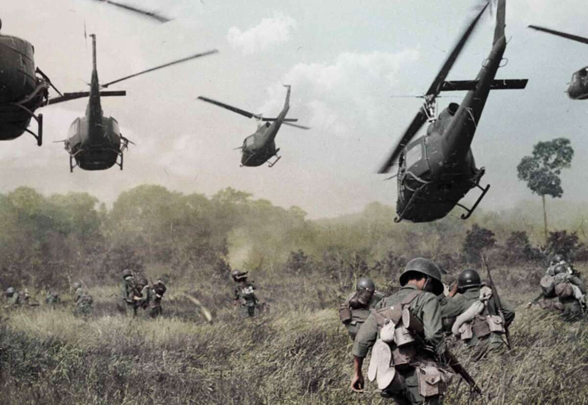 Soldiers in combat gear advance through tall grass as several military helicopters hover above, preparing to land in a rural area with trees and cloudy skies in the background.
