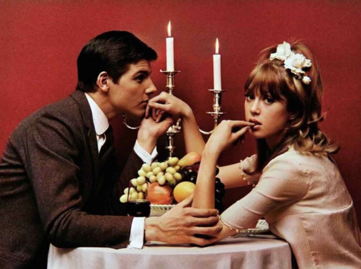 A man in a suit and a woman with a flower headband sit at a table with fruit, holding hands and looking pensive. Two lit candles in a silver holder are in the background against a red wall.