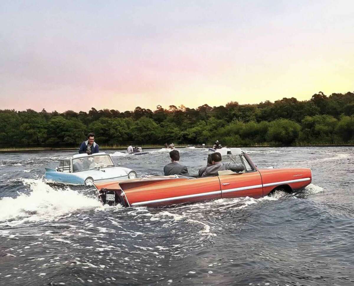 Two classic cars, including a red convertible, drive on a river like boats, with people inside wearing suits. Trees line the riverbank under a colorful sunset sky.