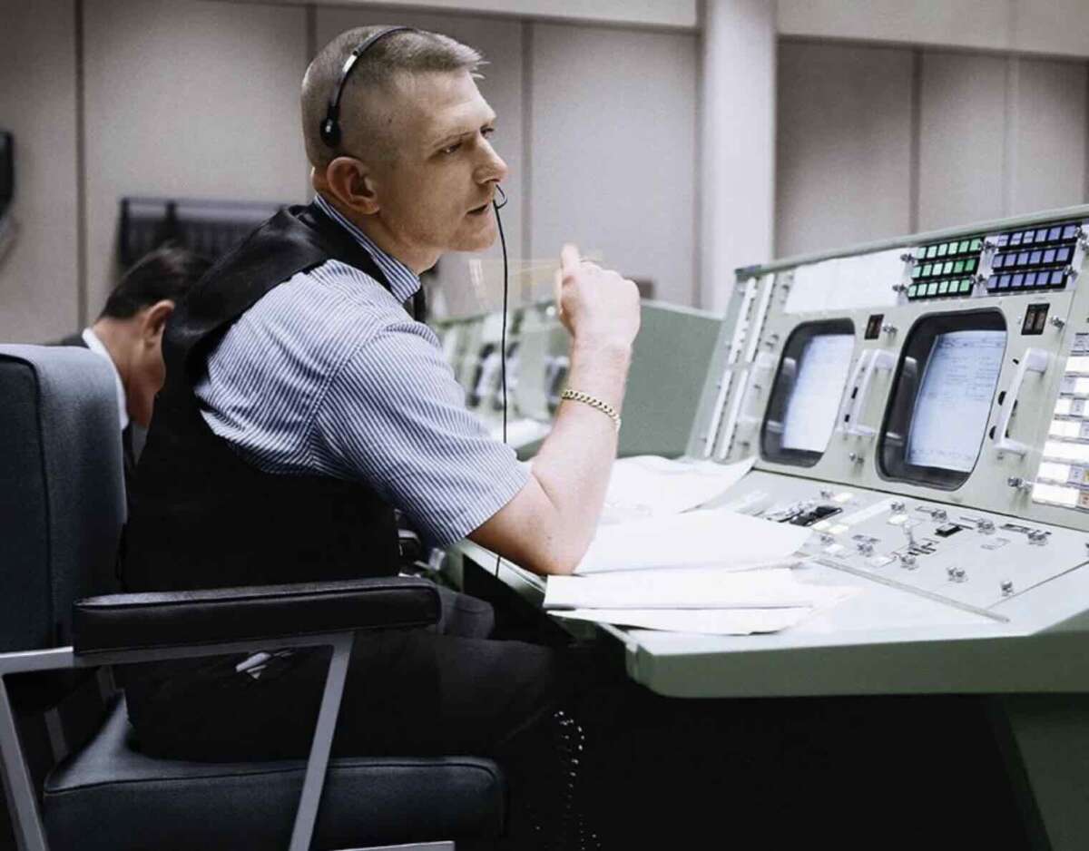 A man wearing a headset and a striped shirt sits at a control panel filled with buttons, dials, and screens in a mission control room, appearing focused and engaged in conversation.