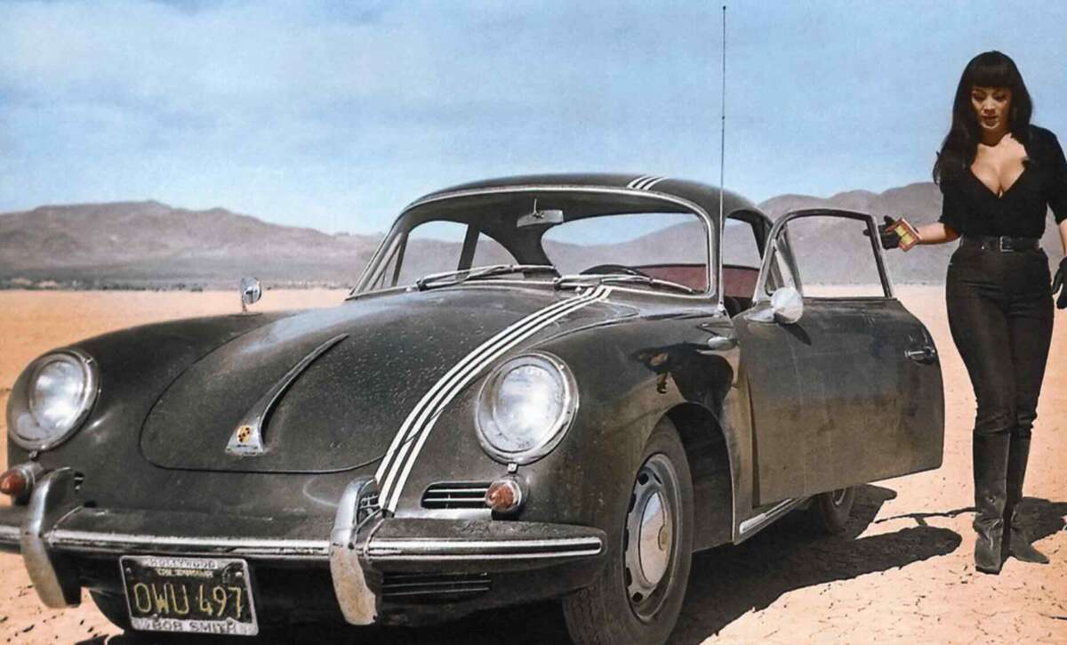 A woman in black clothing stands next to an open car door of a vintage black Porsche, parked on a barren desert landscape with mountains in the background under a blue sky.