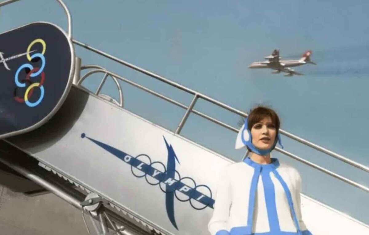 A woman in a retro blue and white flight attendant uniform stands beside airplane stairs marked "Olympic," with an airplane flying in the sky behind her.