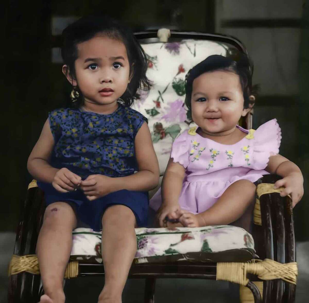 Two young children sit on a cushioned wicker chair; the older child wears a blue outfit and looks slightly to the side, while the younger child in a pink dress smiles at the camera.
