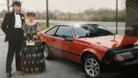 A young couple dressed in formal attire stands by a shiny red sports car, possibly from the 1980s, parked on a street with trees and grass in the background.