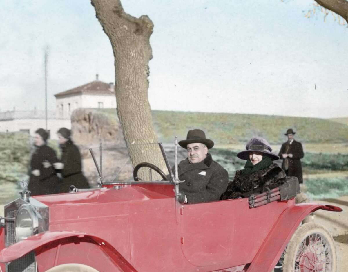 A man and a woman, both wearing coats and hats, sit in a vintage red convertible car parked by a tree. Blurred figures and a house are visible in the grassy background.