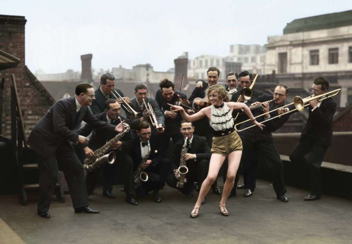 A woman in a striped top and gold shorts dances energetically on a rooftop, surrounded by a lively jazz band of men playing brass and woodwind instruments. The scene appears vintage and joyful.
