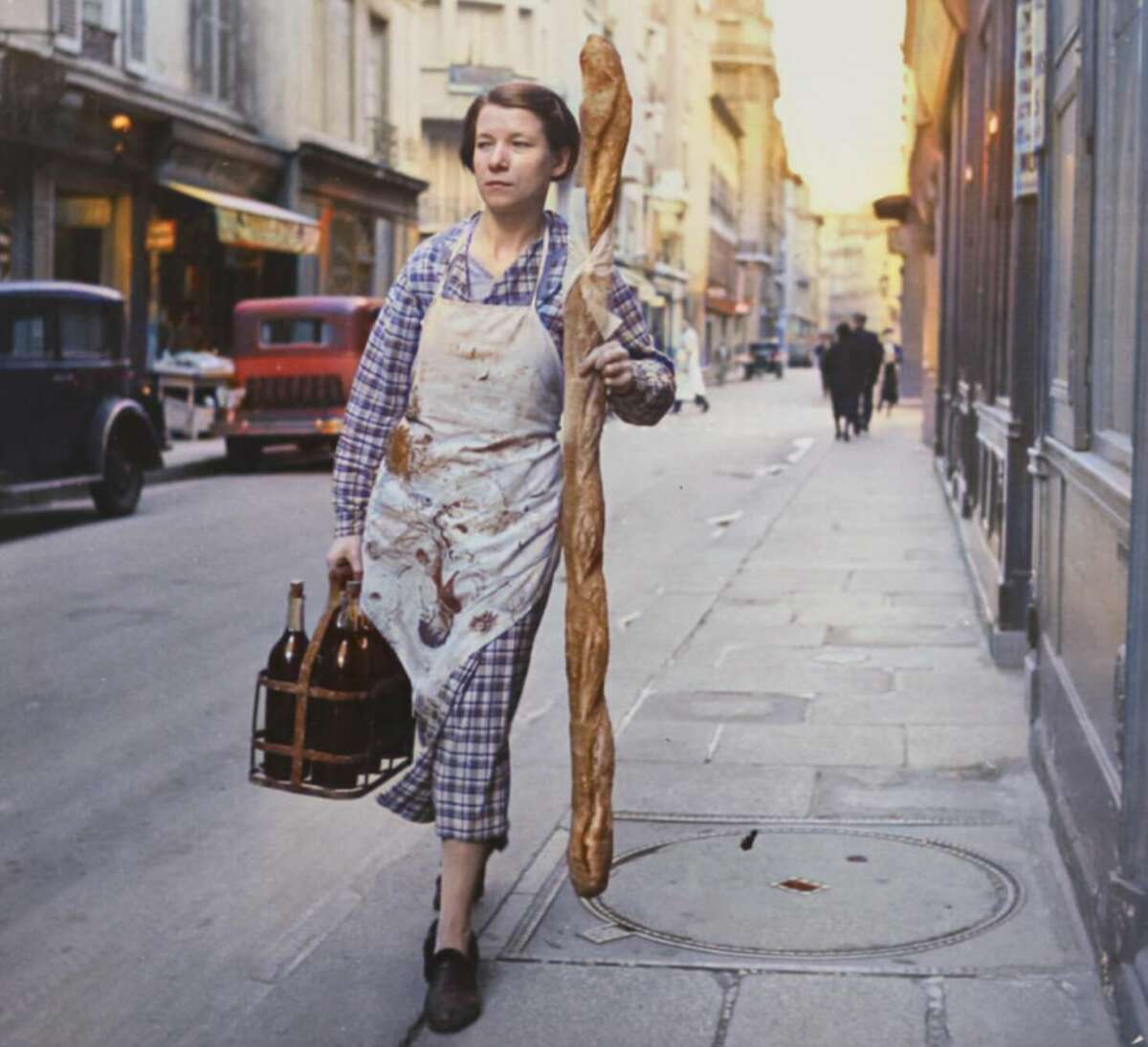 A woman in a checkered dress and stained apron walks down a city street carrying a large baguette and a crate with bottles, with vintage cars and buildings in the background.
