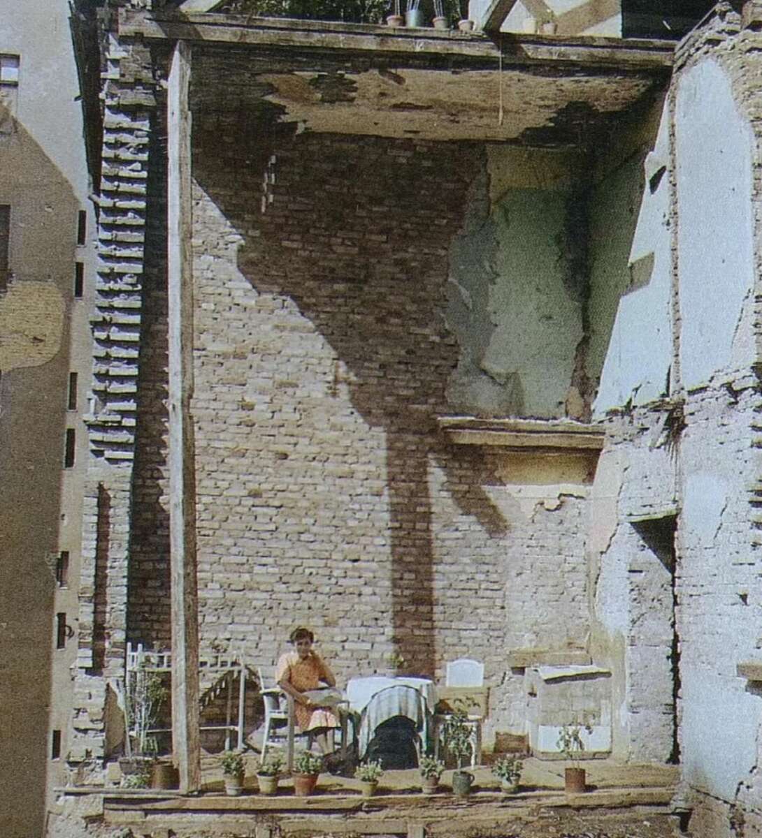 A person sits on a chair in a partially demolished building, surrounded by potted plants and furniture, with exposed brick walls and remnants of rooms visible.