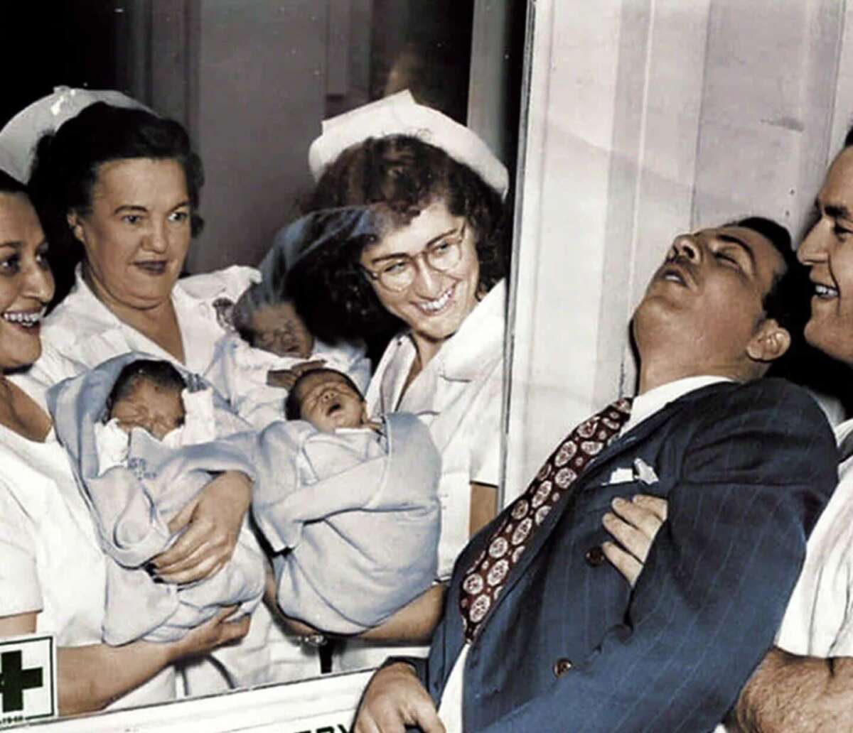 Three nurses hold newborn babies and smile, while a man in a suit appears to faint or swoon, supported by another man, possibly reacting to the sight of the babies through a hospital window.