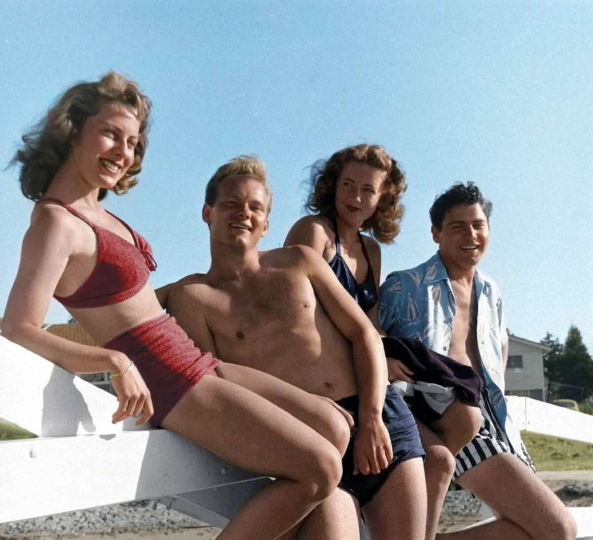 Four young adults in vintage swimwear smile and pose outdoors on a sunny day, sitting casually on a white fence with blue sky and grassy scenery in the background.